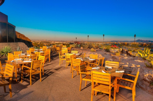 Outdoor restaurant patio with wooden tables and chairs, overlooking a scenic landscape under a clear blue sky.