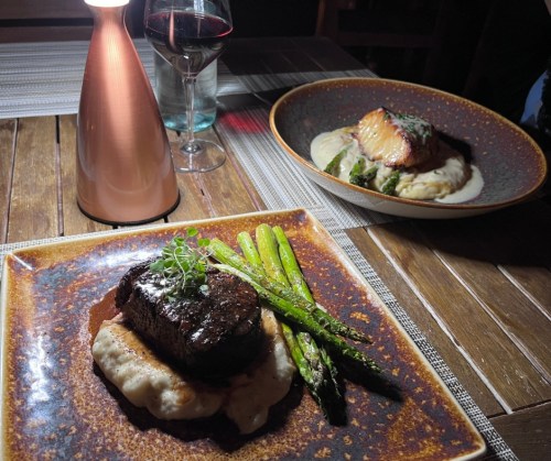 A beautifully plated steak with asparagus and mashed potatoes, alongside a dish of salmon and vegetables, with wine in the background.