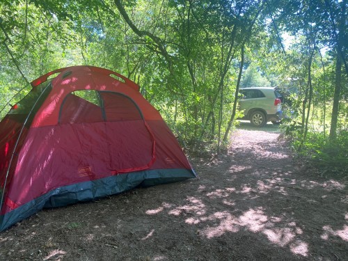 A red tent set up in a wooded area, with a vehicle parked nearby on a dirt path. Sunlight filters through the trees.