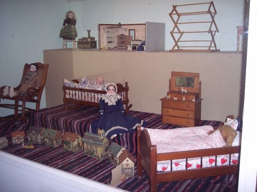 A display of vintage dolls in beds, with a miniature house and furniture in a cozy, colorful setting.
