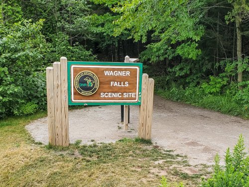 Sign for Wagner Falls Scenic Site, surrounded by greenery and a dirt path leading into the woods.