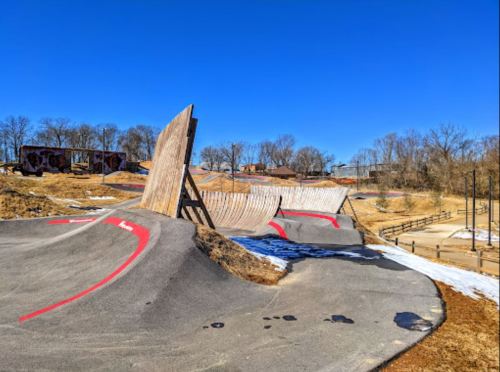 A skate park with a wooden ramp and smooth concrete surface under a clear blue sky. Trees in the background.