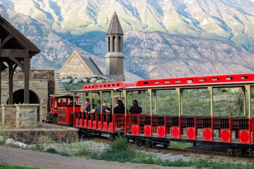 A red train with passengers travels through a scenic landscape with mountains in the background.