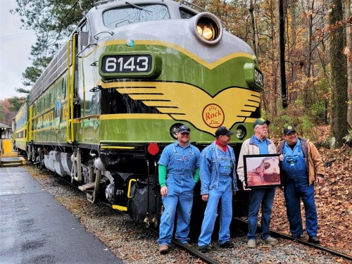 A vintage train with four men in overalls posing beside it, surrounded by autumn foliage.