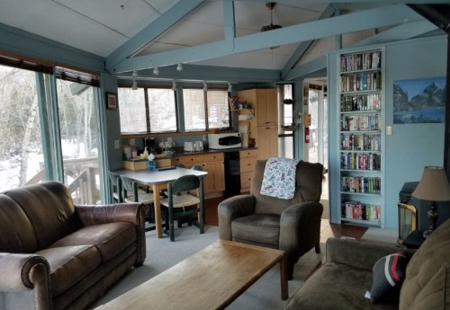 Cozy living area with two sofas, a wooden coffee table, and a kitchen in the background, surrounded by large windows.