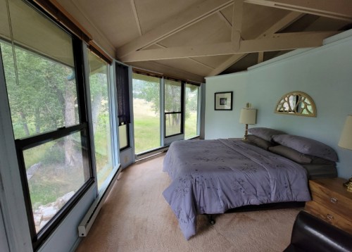 Cozy bedroom with a large bed, soft blue walls, and windows offering a view of greenery outside.