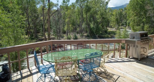 A wooden deck with a round glass table and colorful chairs, overlooking a serene river and lush green trees.