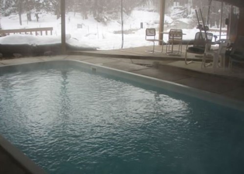 Indoor swimming pool with steam rising, surrounded by snowy landscape and large windows.
