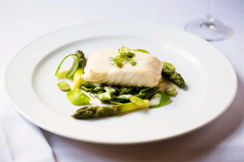 A plate of white fish atop a bed of asparagus and green vegetables, garnished with microgreens.