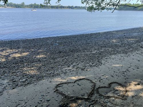 Two heart shapes drawn in the sand on a pebbly beach, with a calm river and trees in the background.