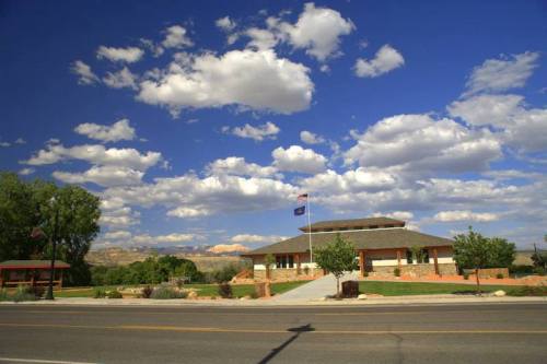 A building with a flag in front, surrounded by trees and a clear blue sky with fluffy clouds.