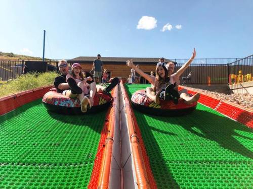 Four children on inflatable tubes slide down a colorful, green slope on a sunny day, smiling and having fun.