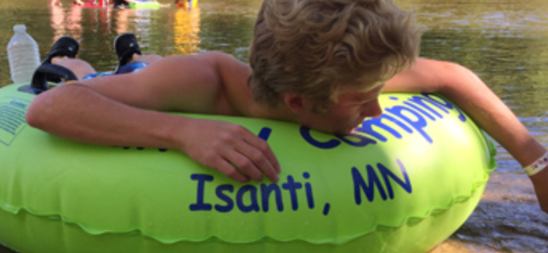 A person relaxes on a bright green inner tube in a river, with a water bottle nearby and others in the background.