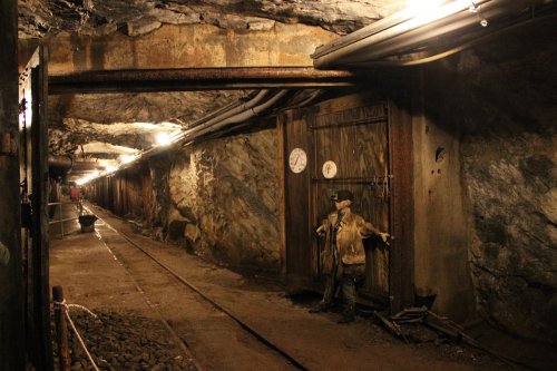 A dimly lit mine tunnel with wooden doors, tracks, and a miner statue standing against the wall.