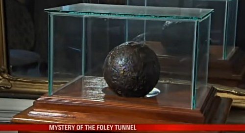 A dark, spherical object displayed in a glass case on a wooden pedestal, titled "Mystery of the Foley Tunnel."