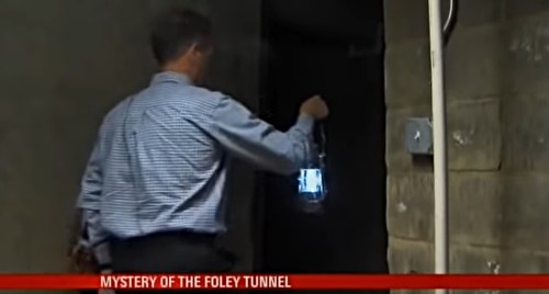 A man holding a lantern stands at the entrance of a dark tunnel, exploring the mystery of the Foley Tunnel.