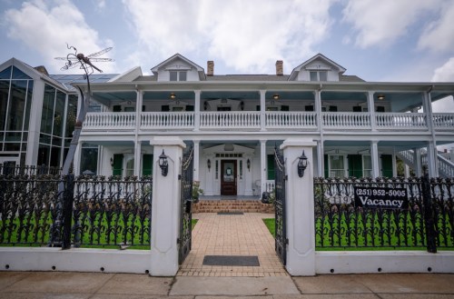 A large, historic house with a porch, surrounded by a black iron fence, featuring a "Vacancy" sign.