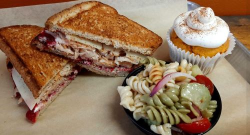 A toasted sandwich with turkey and cranberry, a side of pasta salad, and a cupcake with frosting on a tray.