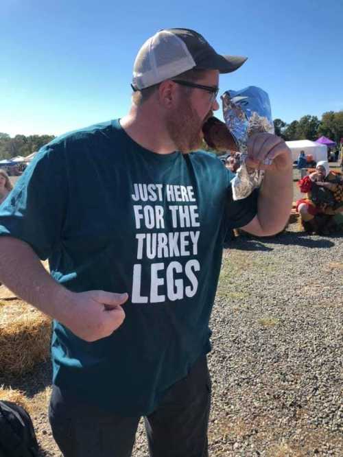 A man in a teal shirt that says "JUST HERE FOR THE TURKEY LEGS" enjoys a turkey leg at an outdoor event.