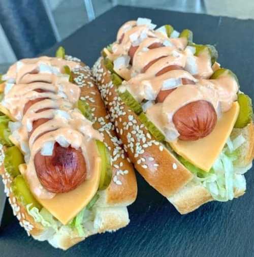 Two hot dogs in sesame seed buns topped with cheese, pickles, onions, and a creamy sauce on a black plate.