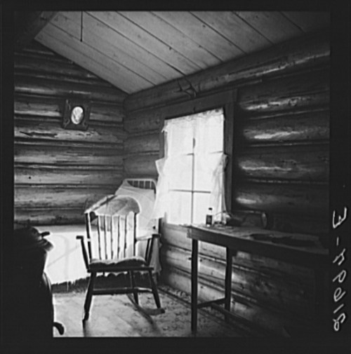 A rustic log cabin interior featuring a wooden chair, a table, and a window with sheer curtains.