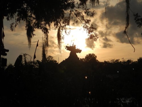 Silhouette of a mountain-like structure against a sunset, framed by trees and clouds.