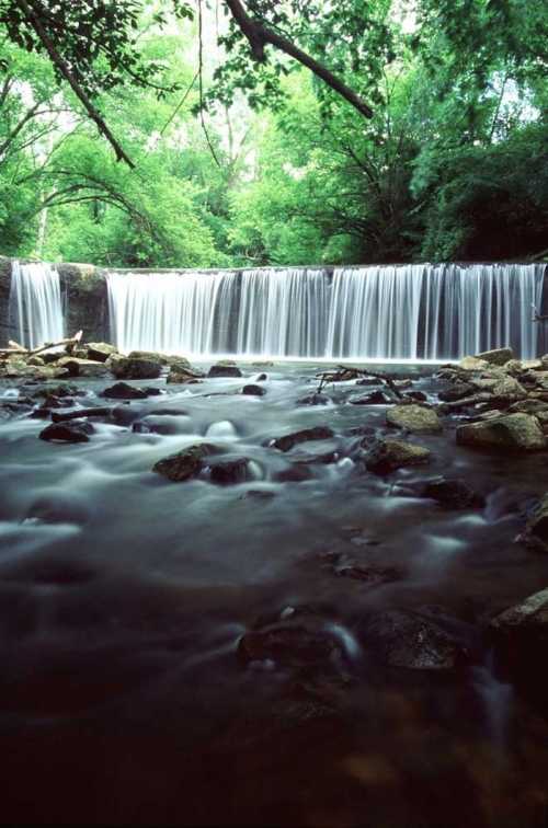 A serene waterfall cascades over rocks, surrounded by lush green trees and a tranquil river.