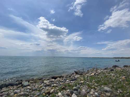 A serene lakeside view with rocky shore, calm water, and a cloudy blue sky.