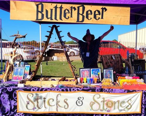 A vendor in a wizard costume stands behind a colorful booth labeled "ButterBeer" and "Sticks & Stones" at a fair.