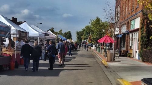 A bustling street market with vendors under tents, people browsing, and shops lining the road on a sunny day.