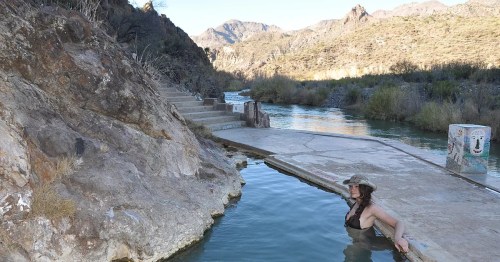 A woman relaxes in a natural hot spring surrounded by rocky terrain and mountains under a clear sky.