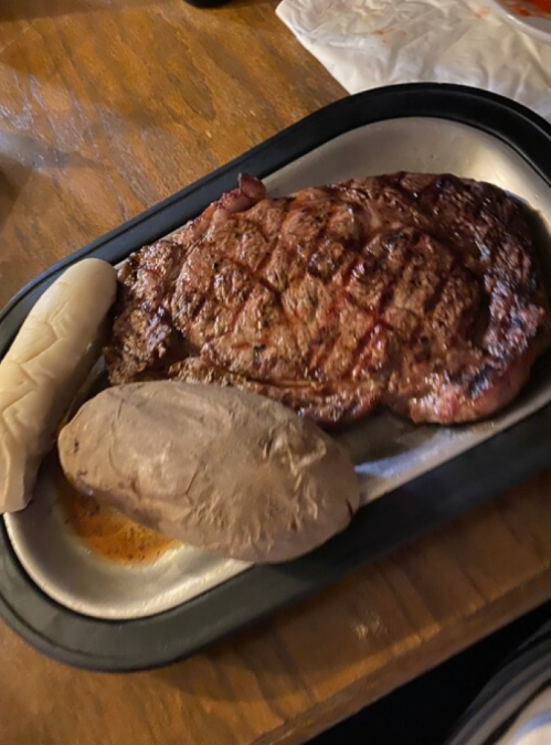 A plate with a grilled steak, a baked potato, and a soft roll on a wooden table.