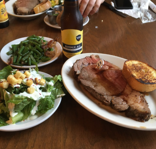 A plate with prime rib, garlic bread, a side salad, green beans, and a bottle of summer ale on a wooden table.