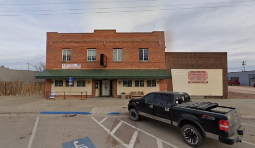 A brick building with a green awning, parked black truck, and signs for a supply store in a small town setting.