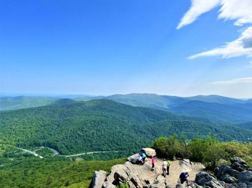A panoramic view from a mountain peak, showcasing lush green hills and a clear blue sky with hikers in the foreground.