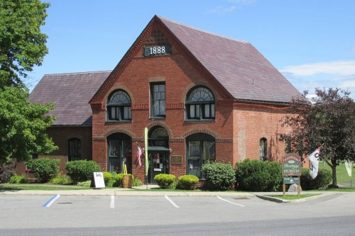 Historic brick building with a steep roof, marked "1888," housing a museum, surrounded by greenery and flags.