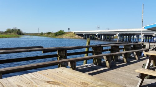 A serene view of a river with a wooden dock, under a clear blue sky and a bridge in the background.