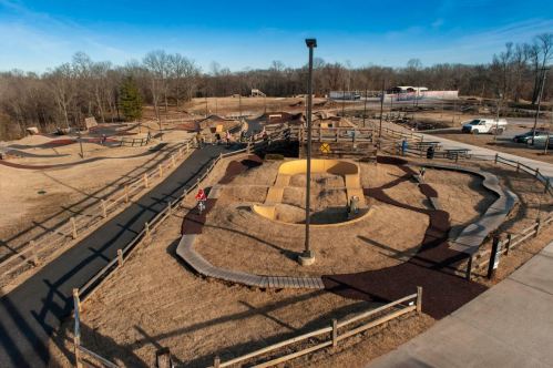 Aerial view of a playground with hills, paths, and wooden fences, set in a grassy area under a clear blue sky.