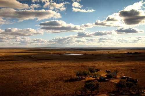 A vast, open landscape with a few scattered houses, under a sky filled with clouds and a distant pond.