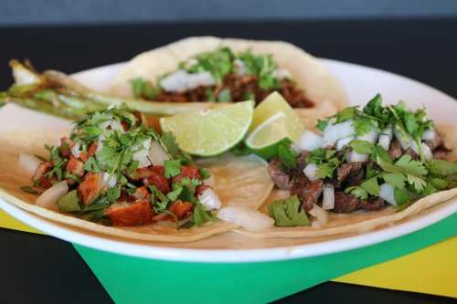 Three tacos on a plate, filled with meat, onions, and cilantro, garnished with lime wedges and grilled peppers.