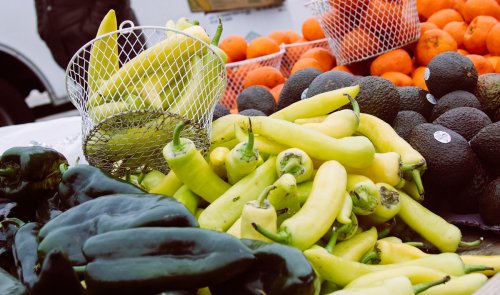 A variety of peppers, including yellow and green, alongside avocados and oranges at a market stall.