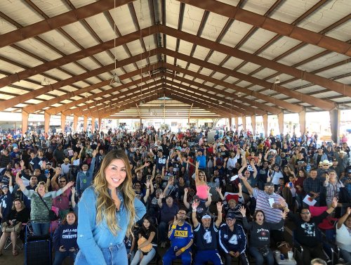 A large crowd of enthusiastic fans gathers under a spacious pavilion, with a woman smiling in the foreground.