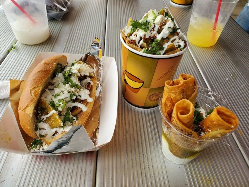 A plate with a sandwich, a cup of loaded fries, and a dessert of rolled pastries, all garnished with toppings.