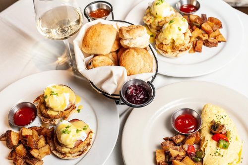 A table setting with two plates of brunch: eggs Benedict, an omelet, crispy potatoes, and a basket of biscuits.