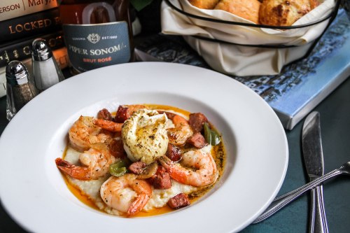 A plate of shrimp and grits topped with sausage, green peppers, and a poached egg, with a drink and bread in the background.