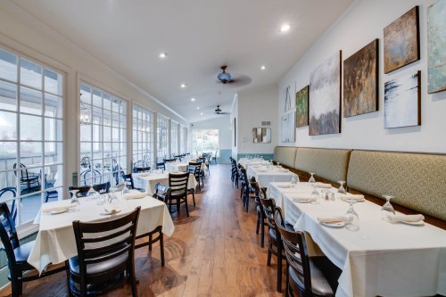 A spacious restaurant interior with white tablecloths, wooden floors, and artwork on the walls, featuring large windows.