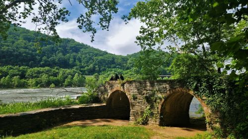 A scenic view of a stone bridge with two arches, surrounded by lush greenery and a river in the background.
