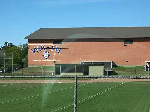 A brick building with "WA-HI" painted on the side, surrounded by green sports fields under a clear blue sky.