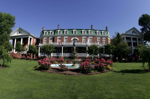 Historic mansion with a green roof, surrounded by gardens and a fountain under a clear blue sky.