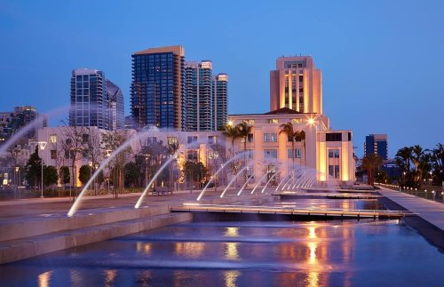 City skyline at dusk with illuminated buildings and a fountain in the foreground.